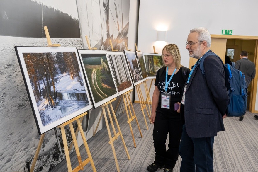 zdjęcie przedstawia dwie osoby oglądające wystawę fotograficzną, przed nimi znajdują się zdjęcia ustawione na sztalugach. Na zdjęciach widoczne są krajobrazy